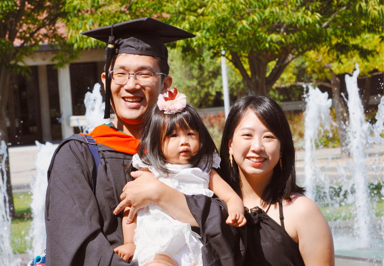 couple holding a child. the man is wearing a graduation gown